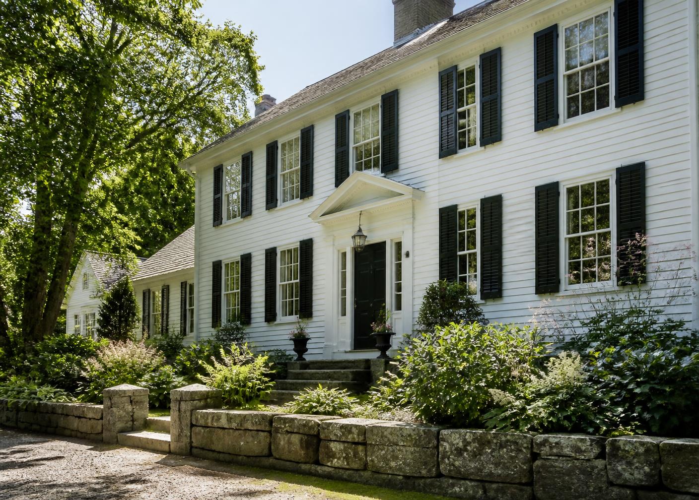 White clapboard colonial home with black shutters, granite steps and stone walls in summer